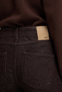 Mörka bordeauxfärgade denimjeans med en texturerad yta, med två bakfickor och en ljus tan läderetikett med "adL" sydd.