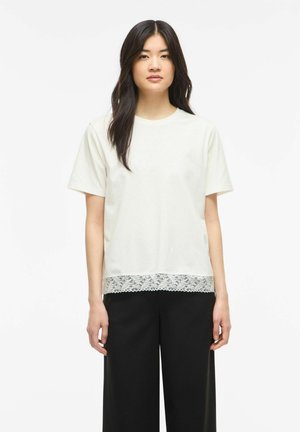 Jeune femme aux longs cheveux foncés portant un haut blanc à manches courtes avec un ourlet en dentelle et un pantalon large noir, debout devant un fond uni.