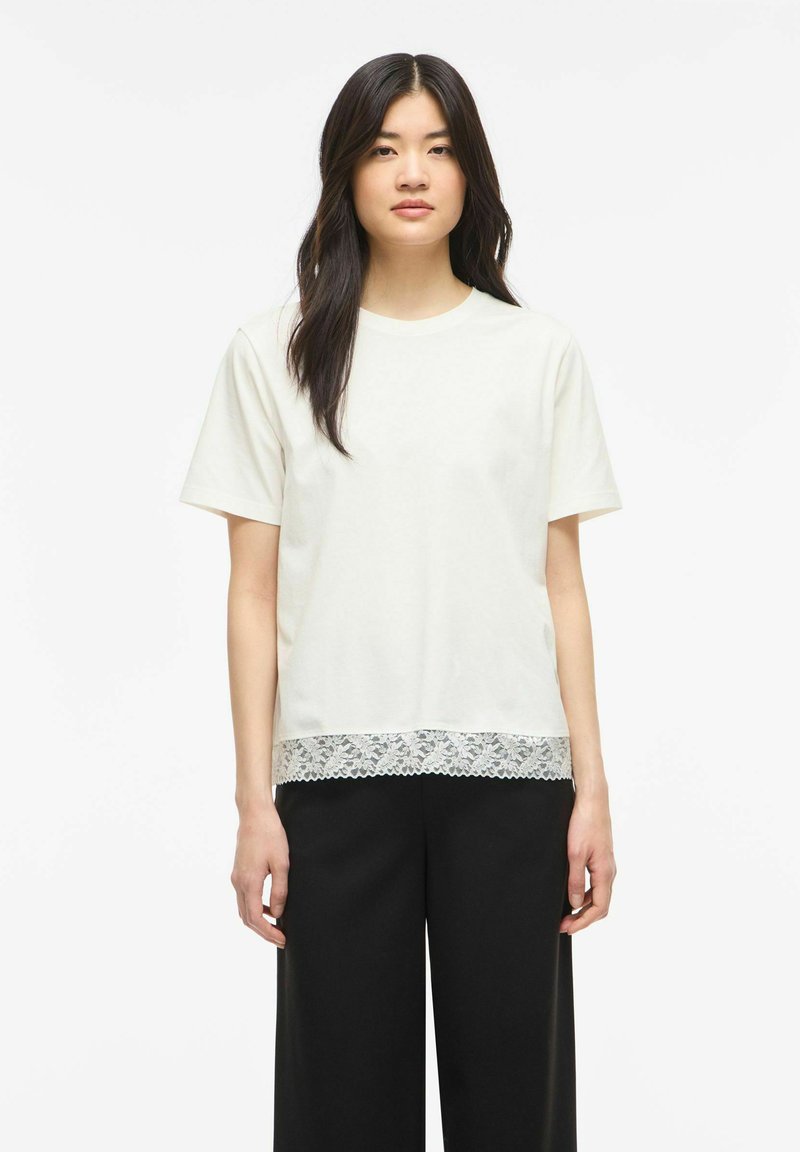 Jeune femme aux longs cheveux foncés portant un haut blanc à manches courtes avec un ourlet en dentelle et un pantalon large noir, debout devant un fond uni.