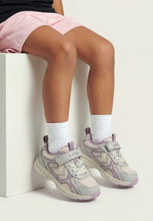 Kind trägt pinke Shorts und weiße Socken, sitzt mit den Füßen in hellgrauen und violetten Sneakers mit Klettverschluss.