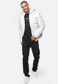 Veste blanche duveteuse avec une surface texturée et une capuche, portée par-dessus une chemise noire, associée à un pantalon noir et des baskets blanches.