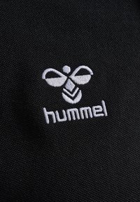 Zwarte stof met een witte geborduurde logo met een bijontwerp boven het woord "hummel." De textuur lijkt glad en dicht geweven.