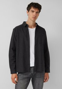 Donkergrijze overhemd met knopen, gemaakt van een gestructureerde stof, met een enkele borstzak en khaki knopen, gedragen over een witte T-shirt.