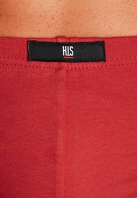 Rotes, tailliertes Kleidungsstück mit einem schwarzen Etikett, auf dem in weißer Schrift "HIS" steht. Das Material wirkt glatt mit einer gerippten Textur.
