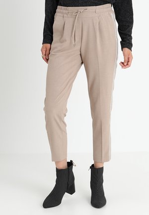 Pantalons beiges à taille élastique avec des bandes latérales, coupe ajustée, en matériau léger. Associés à des bottines noires rayées et un haut sombre.