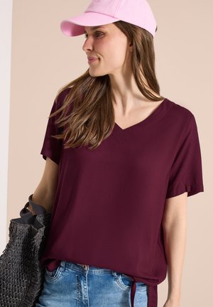 Žena s růžovou čepicí a vínově červenou blůzou s výstřihem do V drží černou pletenou tašku a dívá se stranou před béžovým pozadím.