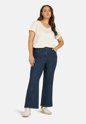 Kvinde med langt hår iført en hvid top med korte ærmer, mørkeblå jeans med vide ben og hvide sneakers, der står med den ene hånd i lommen.