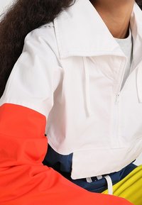 Gekürzte weiße Jacke mit roten Ärmeln, mit einem breiten Kragen und einem Reißverschluss. Das Material wirkt leicht und eignet sich gut für die Freizeit.