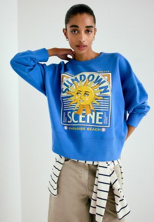 Jauna moteris mėlynu džemperiu su spalvingu saulės grafiku ir užrašu, dėvi smėlio spalvos kelnes ir prie juosmens surištą dryžuotą marškinių uždangalą, pozuoja viduje.