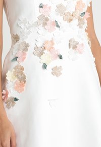 Robe blanche avec des appliqués de fleurs brodés colorés en rose, crème et vert. Texture lisse, corsage ajusté et détails floraux partout.