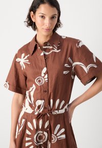 Abito camicia in lino marrone con maniche corte, chiusura con bottoni, motivi astratti bianchi e vita regolabile con cordoncino.