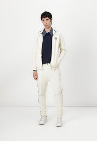 Kapuzenanzug aus Baumwolle in Creme mit marineblauen Akzenten, bestehend aus einer Reißverschlussjacke und gerippten Hosen, kombiniert mit weißen Sneakers.