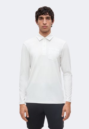 Bărbat purtând un tricou polo alb cu mânecă lungă și buzunar la piept și pantaloni scurți negri, stând în fața unui fundal deschis și simplu.