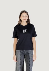 Camiseta de algodón negra de manga corta, con un logotipo estilizado en blanco en el pecho. Combinada con pantalones de mezclilla grises de tiro alto.