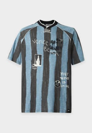 Mann trägt ein blau-schwarz gestreiftes Kurzarmshirt mit Schriftzug und cremefarbene weite Hosen, steht mit den Händen in den Taschen vor einem schlichten Hintergrund.