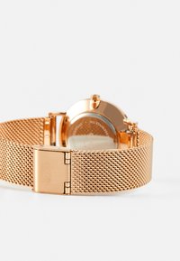Montre en maille rose gold avec un boîtier rond, un bracelet en métal texturé et une fermeture à cliquet. Le dos est visible avec des détails gravés.