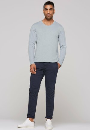 Hellblaues Langarm-V-Neck-Shirt kombiniert mit dunkelblauen Hosen und weißen Sneakern. Der Stoff wirkt glatt und der Schnitt ist locker.