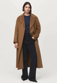 Langer brauner Wollmantel mit Reverskragen, getragen über einem dunkelblauen Oberteil und dunklen, ausladenden Jeans. Glatte schwarze spitze Schuhe runden das Outfit ab.