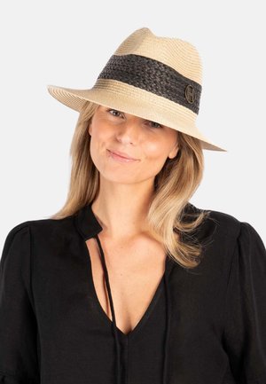 Kvinne med blondt hår iført en beige stråhatt med svart bånd og en svart bluse, smilende lett mot en ensfarget bakgrunn.