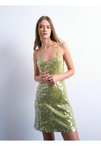 Femme aux longs cheveux portant une robe verte à paillettes sans manches, debout devant un fond clair uni, les mains jointes devant elle.