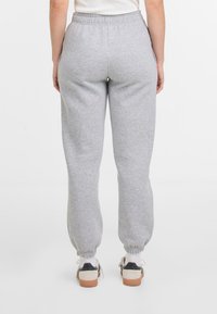 Personne portant un pantalon de survêtement gris clair et des baskets blanches, vue de dos sur un fond blanc uni.