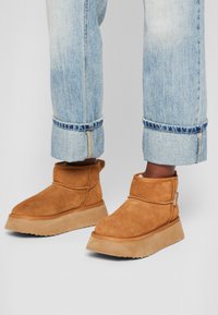 Botine din suede maro cu o talpă plată, texturată și detalii cusute. Designul prezintă un tiv pliabil și un mic mâner pentru a le îmbrăca.
