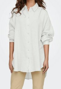 Woman wearing a loose, long-sleeve white button-up shirt with cuffed sleeves and beige pants against a plain background.