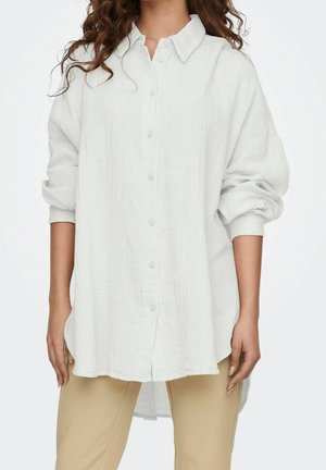 Woman wearing a loose, long-sleeve white button-up shirt with cuffed sleeves and beige pants against a plain background.