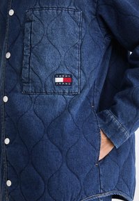 Denimjakke med en quiltet tekstur, mørk blå farge, trykknapper, sidelommer og en Tommy Jeans logo-patch på brystlommen.