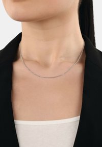 Collana d'argento indossata attorno al collo di una donna sopra una maglietta bianca con un blazer nero, primo piano della clavicola e della parte inferiore del viso.