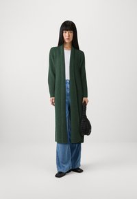 Cardigan lungo verde a costine con apertura frontale, abbinato a una maglia bianca e jeans a gamba larga blu, mentre tiene una borsa nera texture.