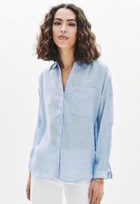 Camicia in lino blu chiaro con chiusura a bottoni, taschino sul petto e maniche arrotolate. Texture morbida e vestibilità rilassata.