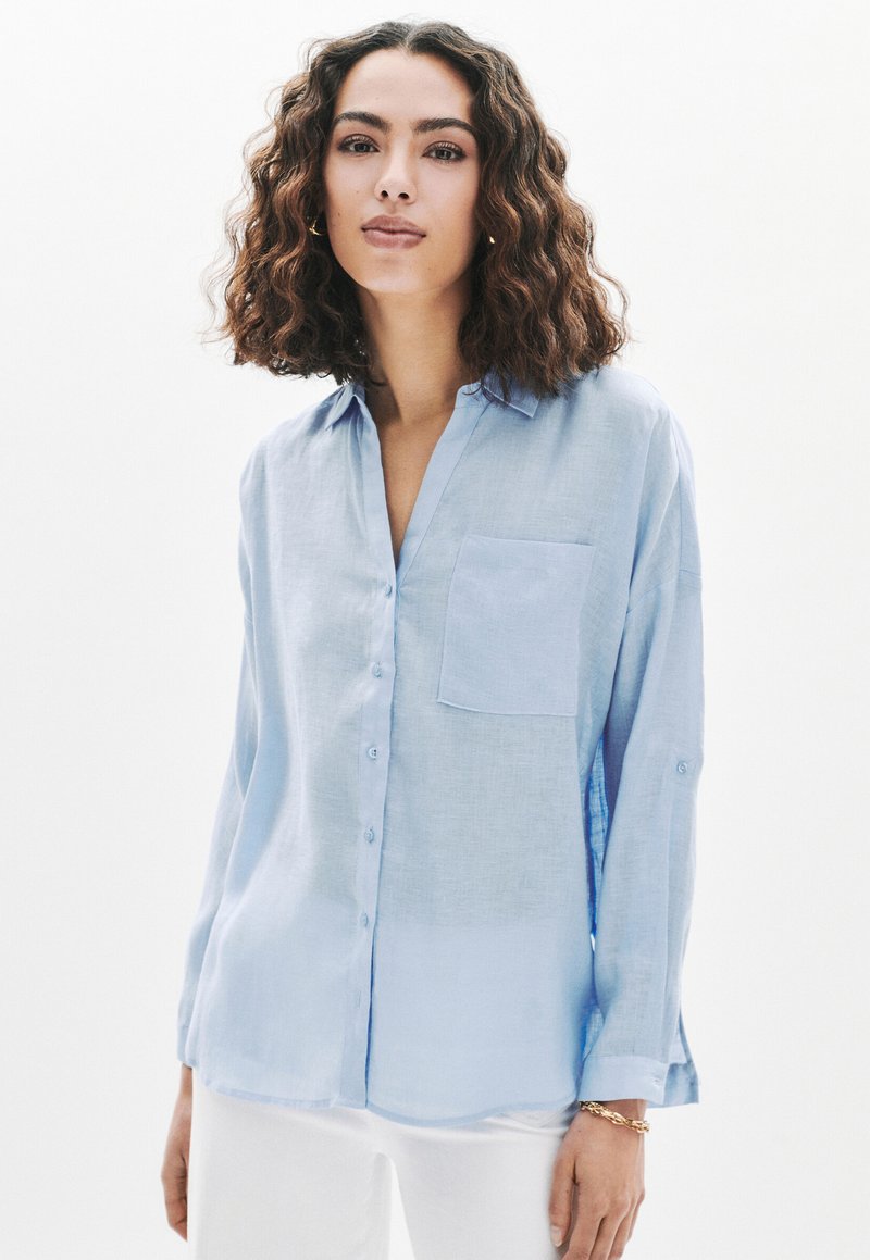 Camicia in lino blu chiaro con chiusura a bottoni, taschino sul petto e maniche arrotolate. Texture morbida e vestibilità rilassata.