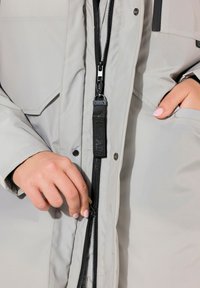 Giacca grigia aderente con una texture liscia, dotata di una chiusura frontale con zip e vestibilità regolabile, accentuata da dettagli in metallo nero e tasche.