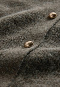 Brunt tekstureret stof med synlige fibre, med en guldfarvet rund knap. Materialet fremstår blødt og let fuzzet.