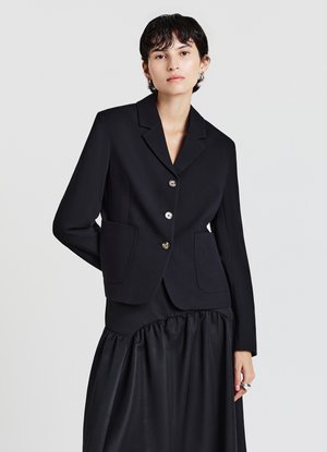 Schwarzer, maßgeschneiderter Blazer mit zwei Fronttaschen, einreihigem Design und metallischen Knöpfen. Getragen über einem schwarzen, gerafften Rock.