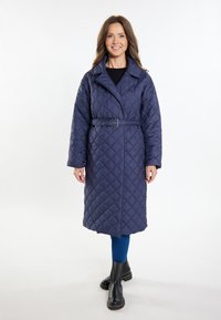 Tengerészkék steppeléses hosszú kabát övvel a deréknál, széles gallérral és ferde elülső zsebekkel. Kék leggings és fekete bokacsizmák fölött viselve.