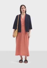 Námornícky prešívaný kabát cez koralovú maxi šaty s gombíkmi a tkanú svetlohnedú crossbody tašku. Tmavé sandále s prekrižovanými pásikmi dotvárajú outfit.