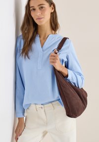 Femme portant une chemise à manches longues bleu clair texturée et un pantalon blanc, tenant un grand sac à bandoulière en daim marron, debout contre un mur.