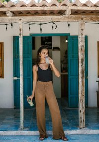 Pantalons en maille marron à jambe large avec une taille à nouer, débardeur noir et sandales marron. Tenant une boisson, debout devant une porte bleue, sur un sol carrelé.