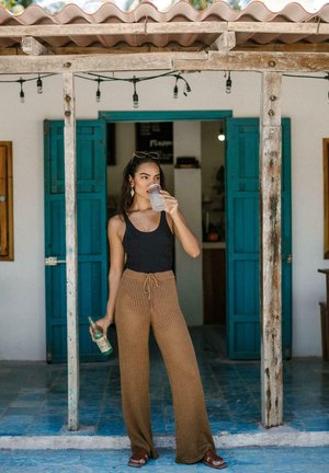 Pantaloni in maglia marrone a gamba larga con vita regolabile, top nero senza maniche e sandali marroni. Tiene una bevanda e si trova davanti a una porta blu, su un pavimento piastrellato.