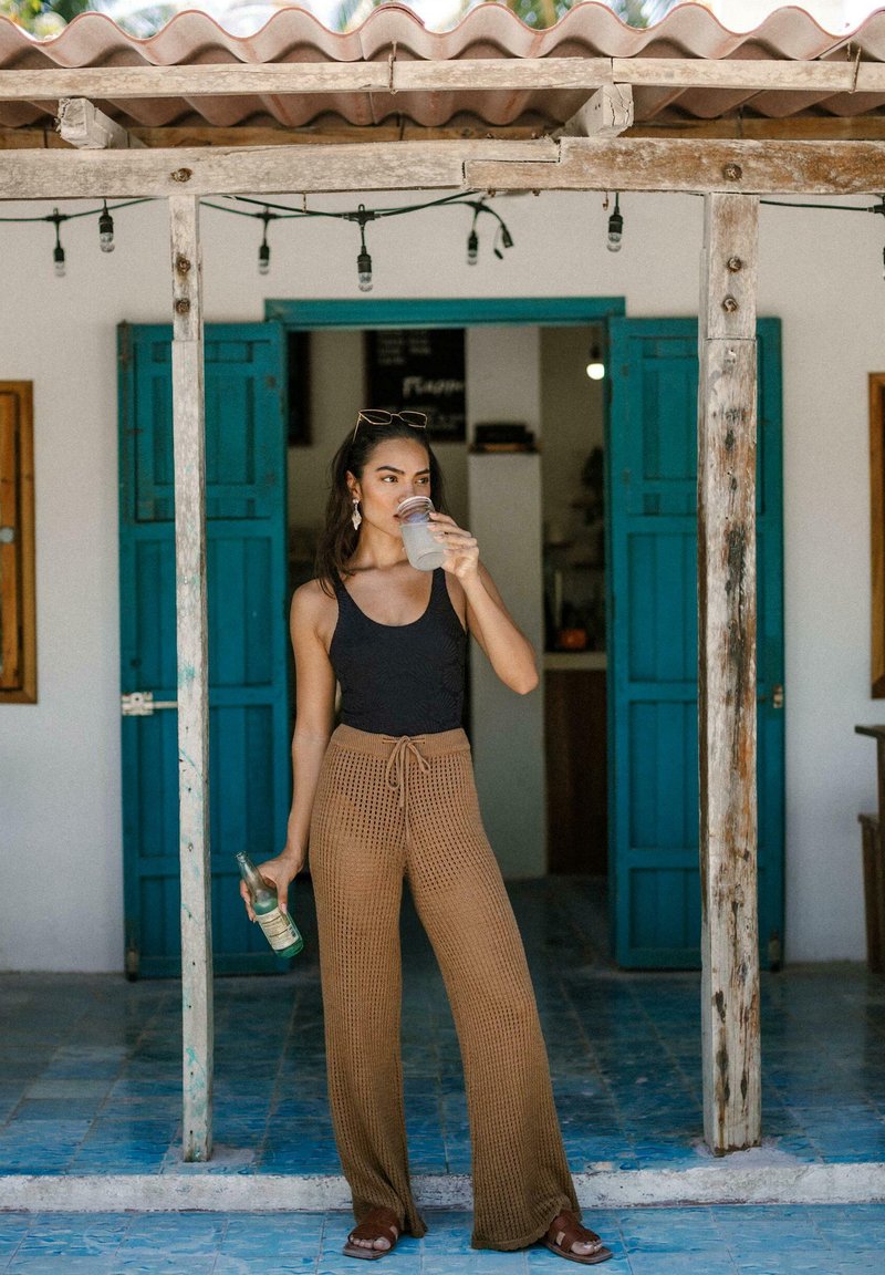 Pantalons en maille marron à jambe large avec une taille à nouer, débardeur noir et sandales marron. Tenant une boisson, debout devant une porte bleue, sur un sol carrelé.