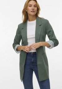 Zielony żakiet o luźnym kroju, z wykładanymi klapami i podwiniętymi rękawami, które odsłaniają paskowane podszewki. Noszony na białej koszuli i niebieskich dżinsach.