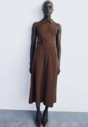 Vestido marrón sin mangas con cuello, botones en la parte delantera y un cinturón en la cintura. Tiene una textura suave y llega por debajo de la rodilla.