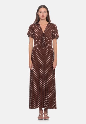 Donna con i capelli lunghi che indossa un vestito maxi marrone con pois bianchi e dettaglio arricciato sul davanti, in piedi davanti a uno sfondo semplice.