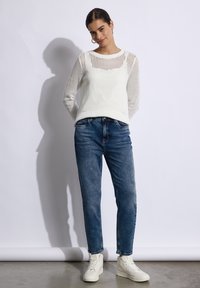 Suéter blanco de punto con un diseño perforado, combinado con jeans de mezclilla azules. El modelo lleva zapatillas altas blancas. El fondo es liso.