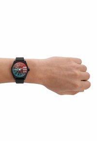 Montre noire hexagonale avec un cadran dégradé rouge et bleu, présentant des marquages blancs, un affichage de la date et un bracelet noir texturé au poignet.