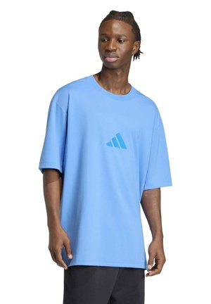 Un mannequin masculin porte un tee-shirt Adidas bleu clair oversize avec un petit logo sur la poitrine et un pantalon noir sur fond blanc.