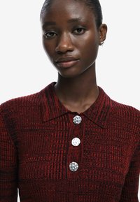 Polo shirt a maglia rossa con texture, colletto e tre bottoni metallici, caratterizzato da un motivo a griglia e una silhouette aderente.