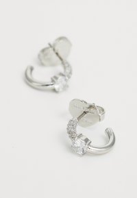 Silver hoop earrings with a curved design, featuring a large clear gemstone and small sparkling accents along one side.